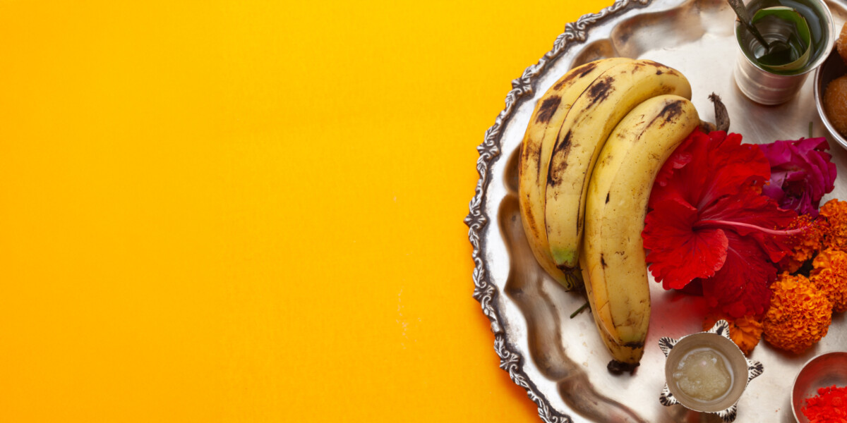 solid yellow background, a silver thali plate on the right with bananas, flowers, paste, diya, silver cup with spoon and water