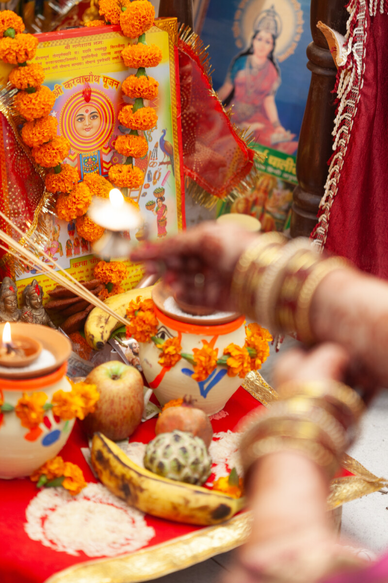 blurred out hands of a Hindu woman performing Aarti to a various deities. Incense, various fruit, diyas, and flowers adorn the image icons.