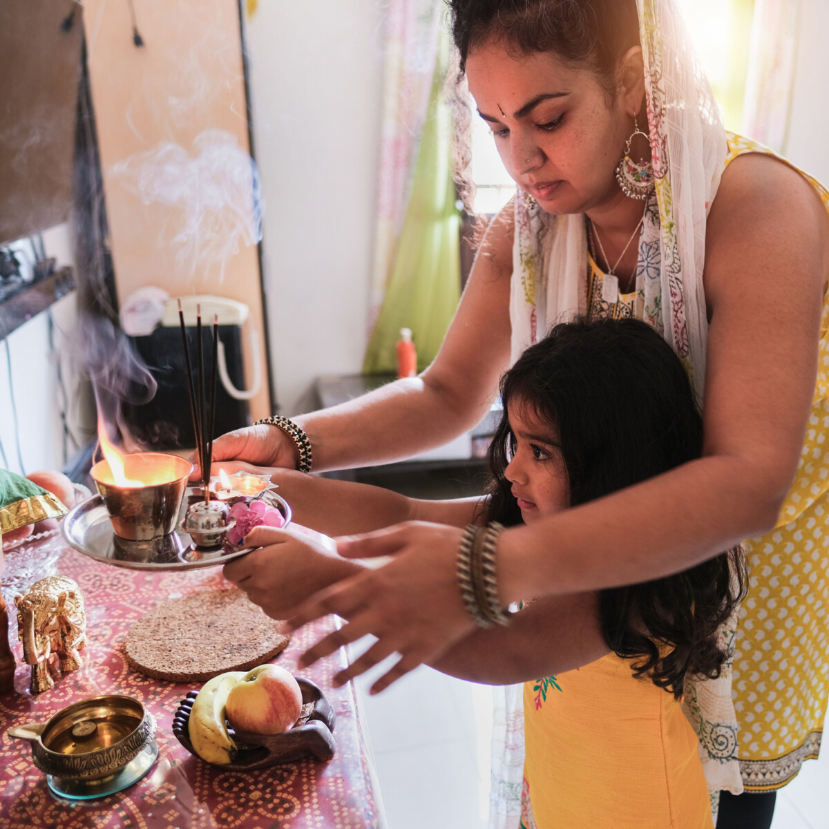 mother with her young daughter on the right, puja items on the left. the mother is guiding her daughter on how to do the puja with the Aarti.