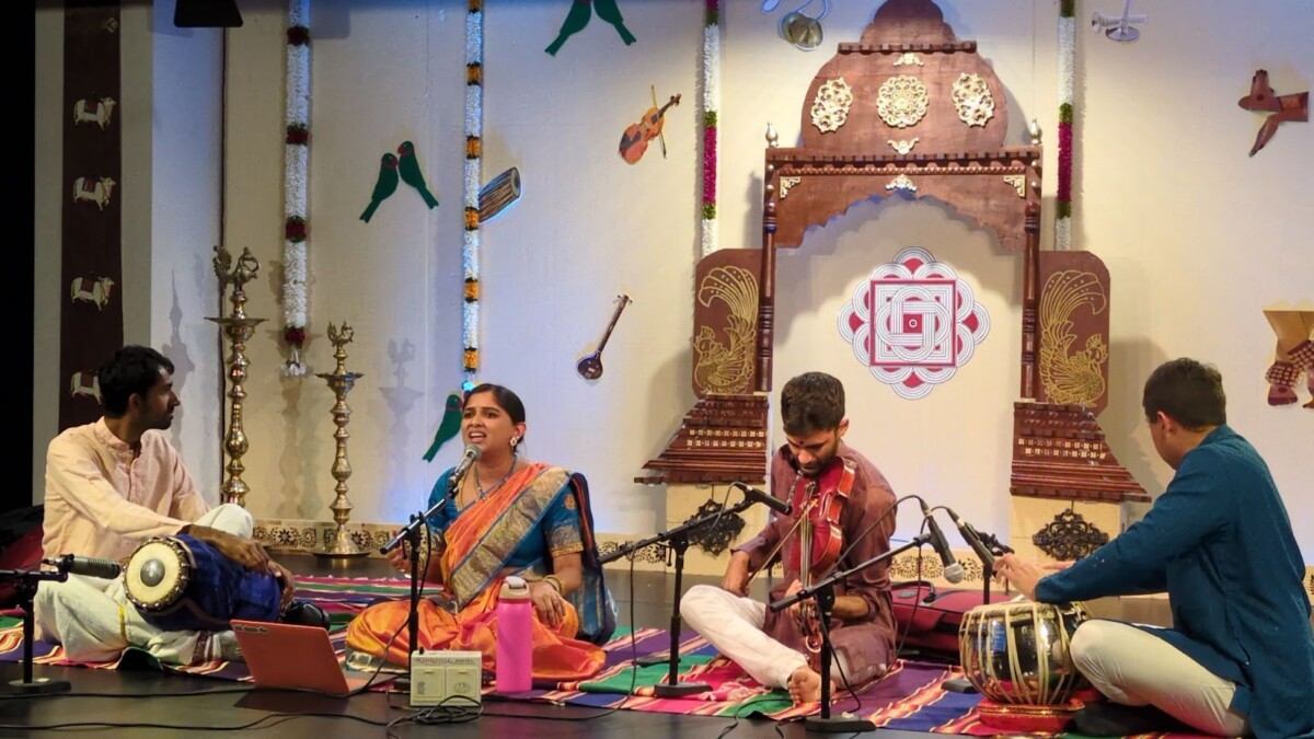 A seated musical performance with a lead lady singer in a sari , surrounded by accompanying male artists playing the violin, mridangam and tabla.