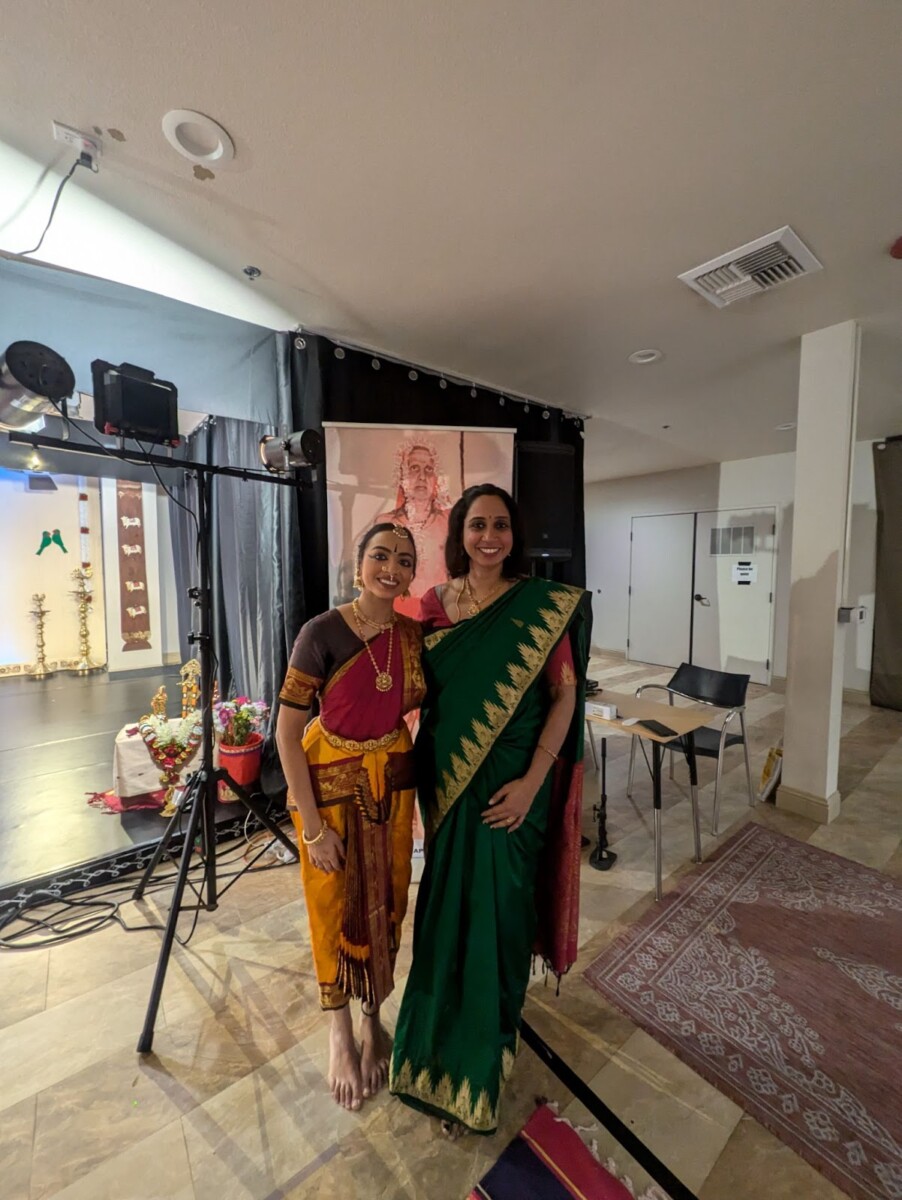 A South Indian classical dancer in traditional costume with a Hindu woman in a sari, in front of the photo of a Hindu saint.