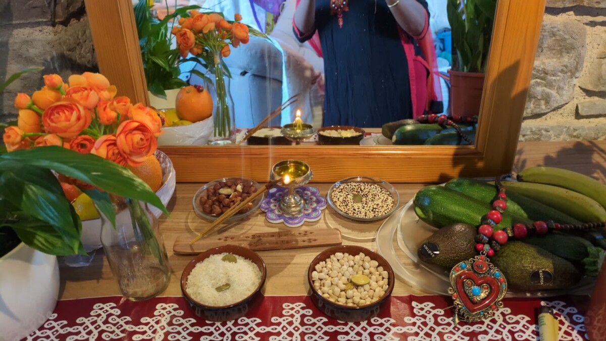 Grains, fruits, flowers and vegetables decorated with jewellery, next to incense and a lighted lamp set in front of a mirror.