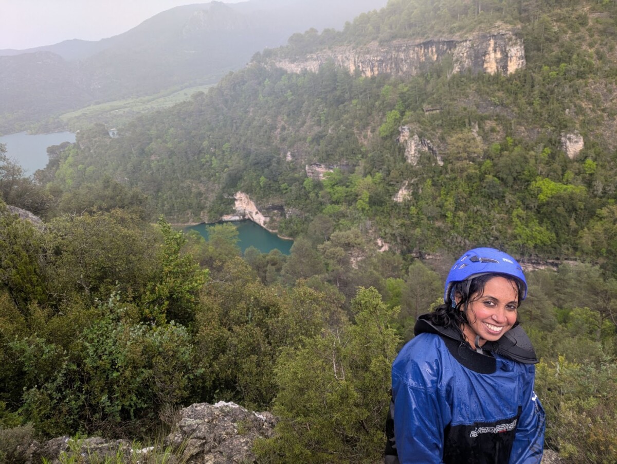 Brown woman in a blue rappelling suit sitting on a rock overlooking a canyon with a river flowing through it.